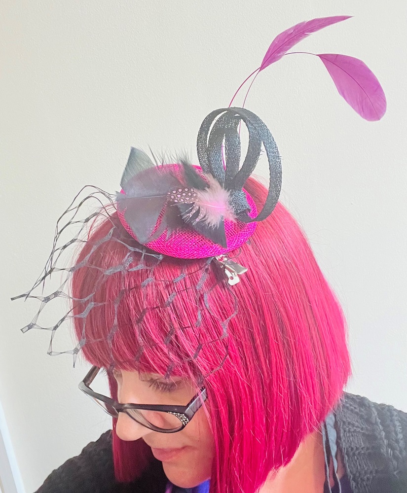 Pink haired woman wearing hand made pink and black fascinator decorated with pink net and feathers in fascinator making workshop.