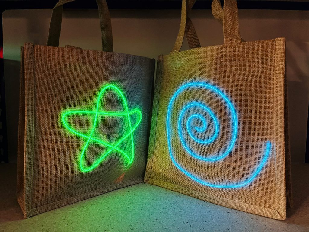 Two hessian tote bags. One has a light up green star design, the other has a light up blue swirl, made in neon workshop.