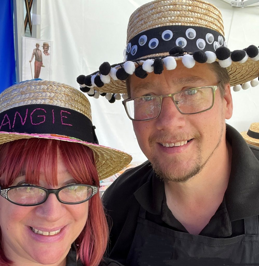 Two people wearing straw boater hats decorated in hat decorating workshop. Man's hat has black ribbon with eyeball details and black and white pompom trim. Woman's hat has black ribbon, pink ricrac and is embroidered "Angie" in pink thread.