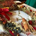 Dried Christmas wreath on table made on straw base ring made with dried green foliage, red flowers, natural wheat and gold berries. Decorated with red bow, pinecones and cinnamon sticks.