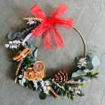 Dried Christmas wreath on wire ring made with dried green foliage and white ruscus, decorated with red bow, white baubles, dried orange slices, pinecones and cinnamon.