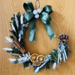 Dried Christmas wreath on wire ring made with dried green foliage and white fluffy pampas grass, decorated with green bow, white baubles, dried orange slices, pinecones and cinnamon.