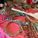 Dried wreath workshop table with straw wreath ring, dried green foliage and dried red flowers on tables. Hands in background making dried Christmas wreaths.