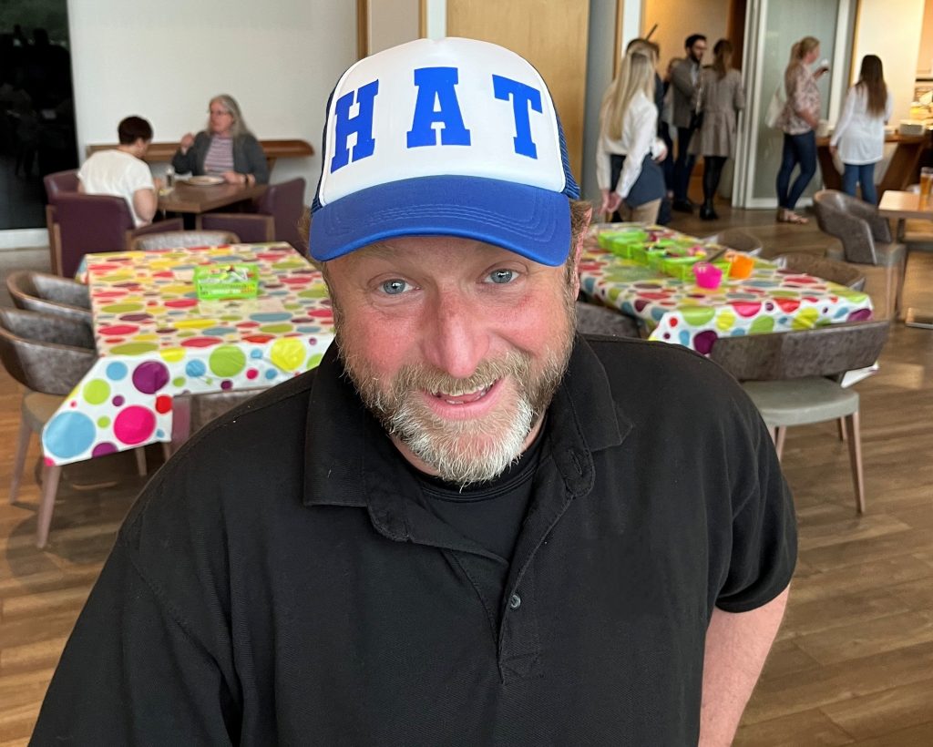 Man wearing blue trucker cap that says "HAT" at a cap decorating workshop.