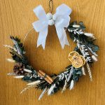 Dried Chritmas wreath on wire ring made with dried green foliage and white fluffy pampas grass, decorated with white bow, white baubles, dried orange slices, pinecones and cinnamon.
