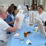 People taking part in corporate painting class. Team painting on canvases on table-top easels. Table set with blue table cover, paints, brushes, palettes and water cups.