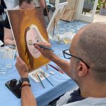 Man painting portrait of a woman on canvas on table top easel during corporate painting class.