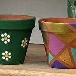 Two hand painted terracotta plant pots painted in plant pot painting workshop. One has green background with white daisies, the other is geometrical design in pink, gold, purple and turquoise.