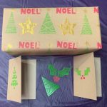 Items made in lino printing workshop - rectangular box wrapped in Christmas printed brown paper with star, tree and "Noel" design, cards printed in holly and tree design, holly and tree lino cut stamps in middle.