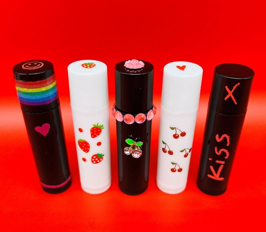 3 black lipstick cases and 2 white lipstick cases, decorated with strawberry and cherry stickers and the word "kiss" to depict a lip balm making workshop.