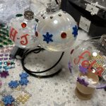 Bauble painting workshop in progress - two glass baubles hang from a bauble hanger, one sits in an egg cup. Stickers are on the table. Baubles are decorates with writing and stickers.