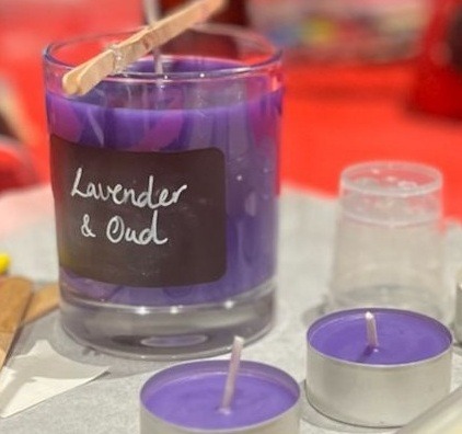 Candle being made in candle making workshop. Purple candle with long wick in wick stabiliser and label that says "Lavender and Oud". Small purple tealights in foreground.