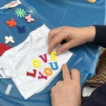 Hands making teddy bear T shirt that says "Love You" made in charity team building activity.