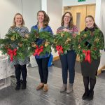 Four women holding up traditional Christmas wreath made with green foliage, pine cones, oranges, cinnamon red baubles and red bow,