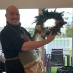 Wreath workshop tutor in florist's apron hold up finished traditional Christmas wreath made with green foliage, pine cones, oranges, cinnamon red baubles and red bow.