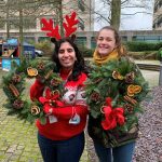 Two women holding up traditional Christmas wreath made with green foliage, pine cones, oranges, cinnamon red baubles and red bow, One woman wears a Christmas jumper and reindeer antlers.