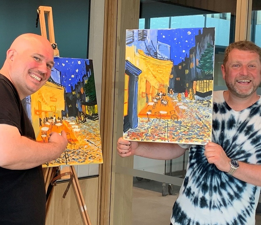 Two men holding paintings similar to Van Gogh's "Cafe Terrace at Midnight" painted in corporate art class.