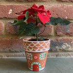 Hand painted terracotta pot painted in gingerbread house design at corporate plant pot painting workshop. Pot holds a poinsettia plant.