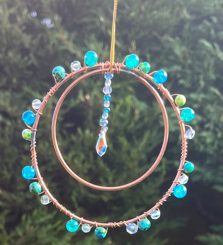 Sparkly suncatcher made in suncatcher making workshop, comprised of two metal rings and decorated with crystals, hanging outside in front of trees with sun shining on crystals.