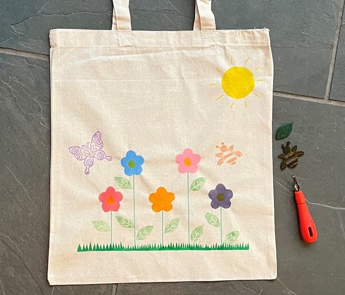 Canvas tote bag featuring flower, butterfly and sun design, with lino printing tool and stamps to the side to depict lino printing workshop.
