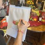 Scene of a watercolour painting class. Hands painting a display of fruit and flowers on an easel.
