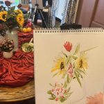 Scene of a watercolour art class. Painting of flowers in watercolour on easel in front of a display of fruit and flowers on a red velvet table cloth.